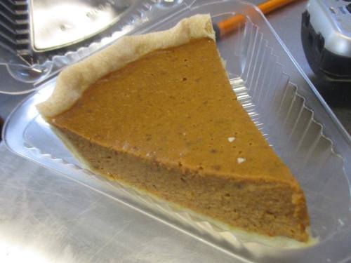 A slice of pumpkin pie in a clear plastic container, showcasing its smooth, spiced filling and golden crust.