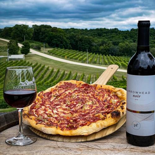 A wooden board with a pizza and a glass of red wine, set against a scenic vineyard landscape under a cloudy sky.