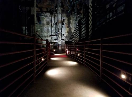 A dimly lit industrial corridor with metal railings and scattered lights, leading into a shadowy space.