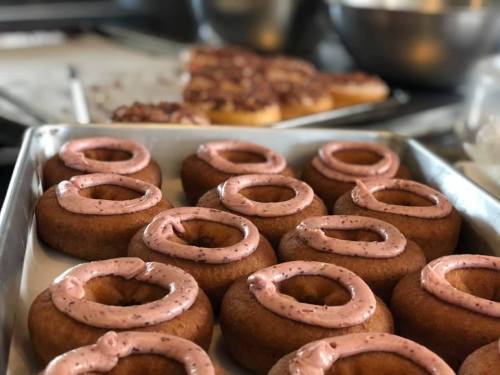 A tray of freshly baked donuts with pink frosting, with more donuts in the background.
