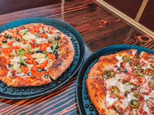 Two pizzas on black trays: one topped with vegetables and olives, the other with meat and jalapeños, on a wooden table.