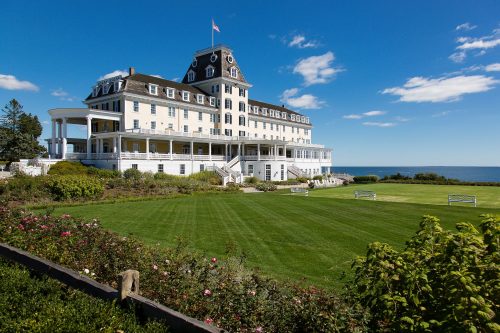 A large historic hotel with a white facade, surrounded by green lawns and blue ocean under a clear sky.