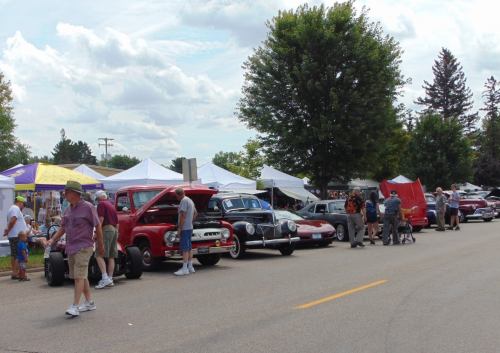 A street lined with classic cars and people browsing tents at an outdoor event on a sunny day.