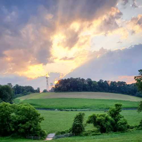 A scenic landscape featuring green fields, trees, and a colorful sunset with clouds in the sky.