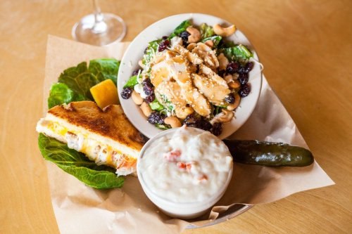 A plate with a grilled sandwich, salad with nuts and berries, and a cup of creamy soup, garnished with a pickle.