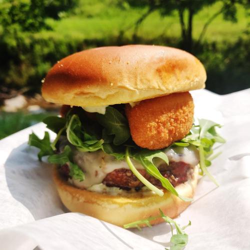 A delicious burger with a golden bun, crispy chicken patty, arugula, and a crispy nugget on top, set against a green background.