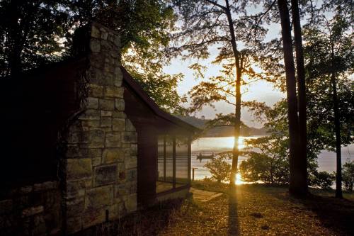 A serene lakeside scene at sunset, featuring a stone cabin and trees reflecting on the water.