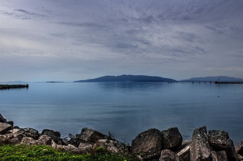 A calm body of water reflects a cloudy sky, with distant islands and rocky shoreline in the foreground.