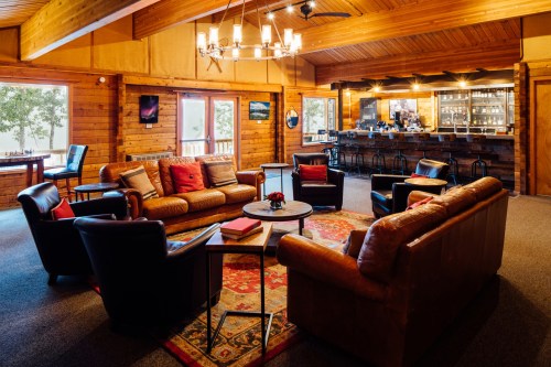 Cozy lounge area with leather sofas, a coffee table, and a bar in a wooden cabin setting, featuring large windows.