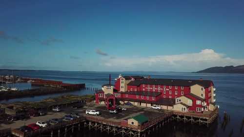 Aerial view of a red and beige waterfront hotel on a pier, surrounded by water and distant hills under a clear blue sky.