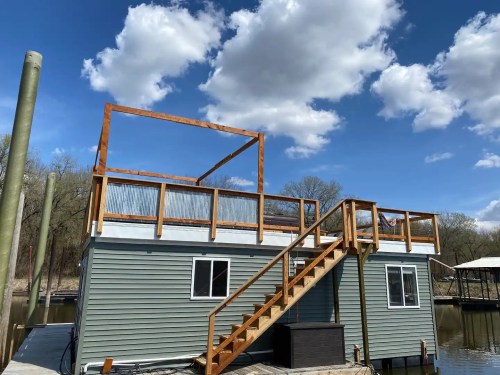 A houseboat with a wooden deck and stairs, set against a blue sky with fluffy clouds.