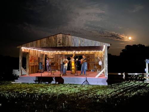 A band performs on an outdoor stage at night, illuminated by string lights and a full moon in the background.