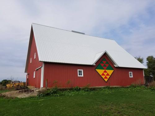 A red barn with a white roof, featuring a colorful geometric quilt pattern on its side, surrounded by green grass.
