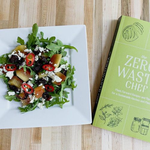 A fresh salad with arugula, cheese, and peppers, next to "The Zero Waste Chef" cookbook on a wooden surface.