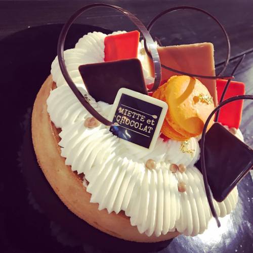 A beautifully decorated cake with whipped cream, chocolate pieces, and a macaroon on top, featuring a decorative sign.