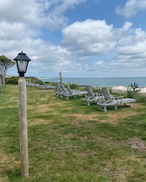 A grassy area with lounge chairs facing the ocean, a lamp post nearby, and a cloudy sky above.