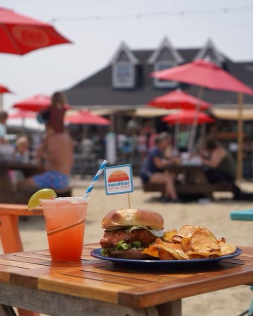 A beachside table with a burger, chips, and a drink, surrounded by umbrellas and people enjoying the sun.