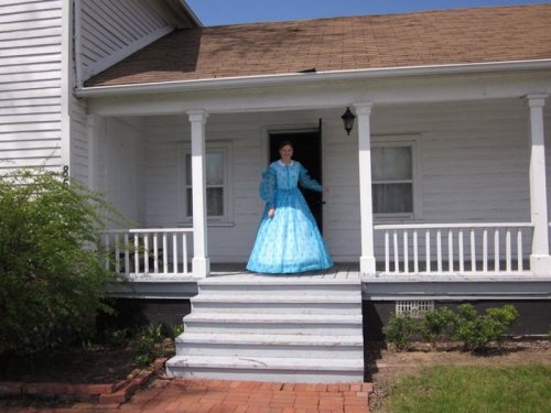 A person in a blue vintage dress stands on the porch of a white house, smiling and welcoming.