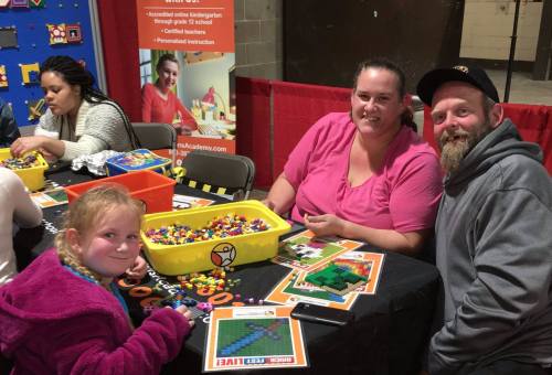 A family enjoys a crafting activity at a table, with colorful beads and projects in a community event setting.