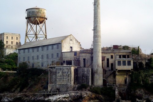 Abandoned industrial buildings with a water tower and smokestack, surrounded by greenery and rocky shoreline.