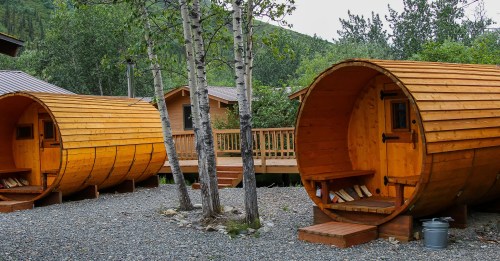 Two wooden barrel-shaped cabins surrounded by trees and gravel, with a deck in the background.