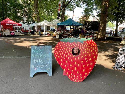 A colorful strawberry cutout with a smiling person inside, next to a sign celebrating Farmers Market Week.