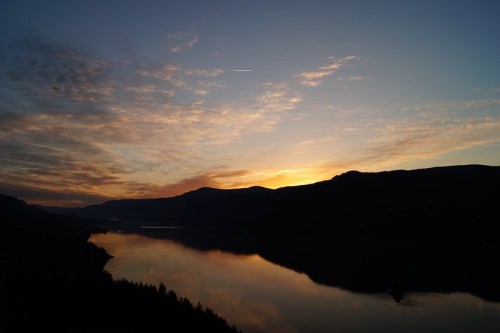 A serene sunset over a calm river, with silhouettes of mountains and colorful clouds reflecting on the water.