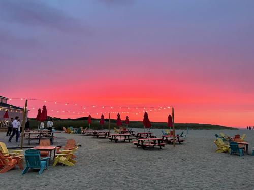 Colorful beach chairs and tables under string lights, with a vibrant pink and orange sunset in the background.