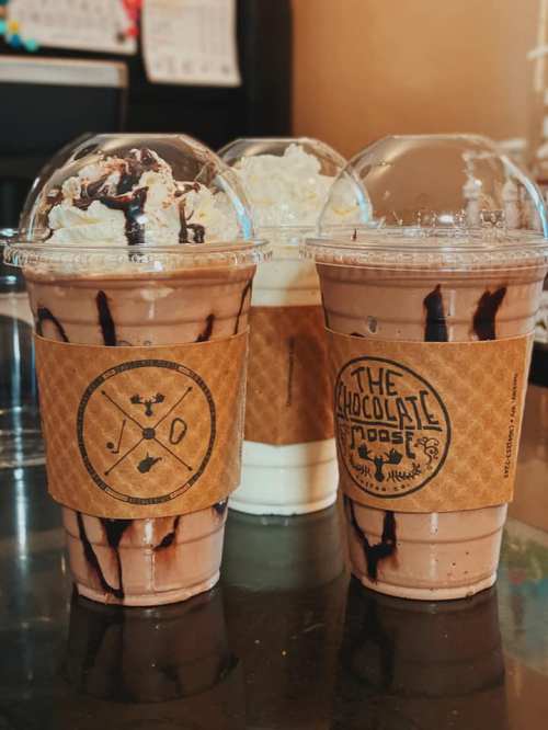 Three milkshake cups with whipped cream and chocolate drizzle, two in front and one behind, on a dark table.