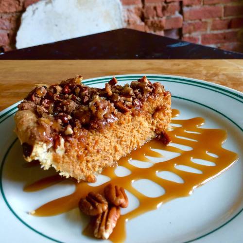 A slice of pecan pie on a plate, drizzled with caramel sauce, with a rustic brick background.