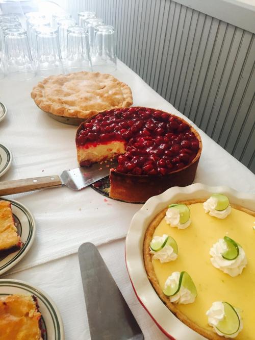 A table with three desserts: a cherry pie, a cheesecake with cherries, and a lemon pie with lime slices and whipped cream.