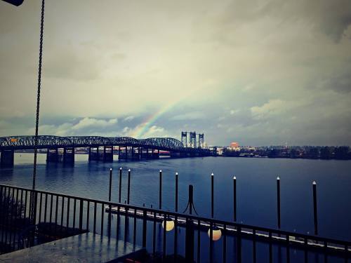 A bridge spans a river under a cloudy sky, with a rainbow arching in the background.