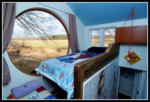 Cozy bedroom with a circular window overlooking a scenic landscape, featuring a wooden bed and colorful bedding.