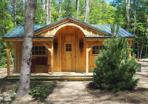 A cozy wooden cabin surrounded by trees, featuring a front porch and a green metal roof.