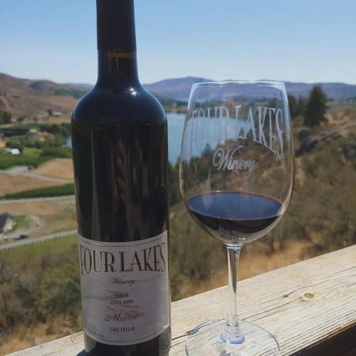 A bottle of Four Lakes Winery Merlot next to a glass of red wine, with a scenic landscape in the background.