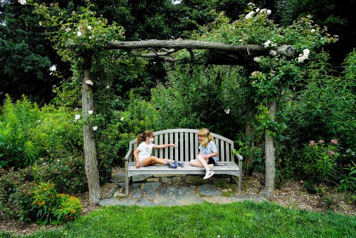 Two children sit on a wooden bench under a floral arch in a lush garden, engaged in conversation.