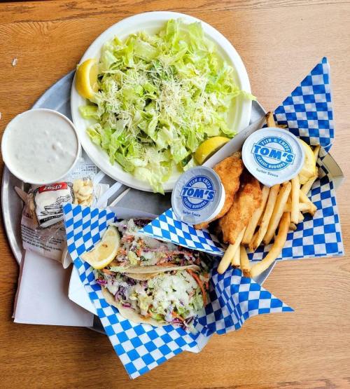 A tray with a salad, fish and fries, and two tacos with slaw, served with lemon wedges and dipping sauces.