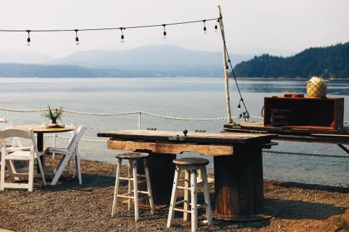 A rustic outdoor dining area by the water, featuring wooden tables, stools, and string lights against a scenic backdrop.