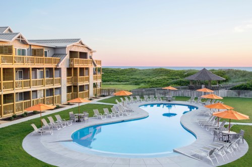 A serene pool area with orange umbrellas, lounge chairs, and a beachfront hotel at sunset.