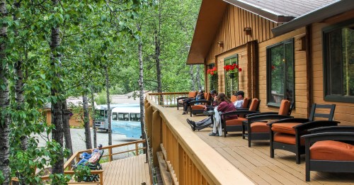 A wooden deck with people relaxing in chairs, surrounded by trees and a vintage bus in the background.