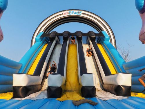 Two children slide down a large inflatable slide on a sunny day, with a blue sky in the background.