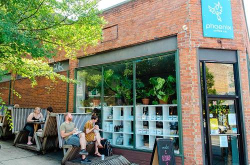 A cozy coffee shop with a brick exterior, featuring outdoor seating and large windows showcasing plants and products inside.