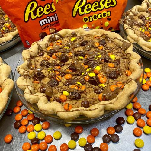 A chocolate pie topped with Reese's Pieces and drizzled with chocolate, surrounded by bags of Reese's candies.