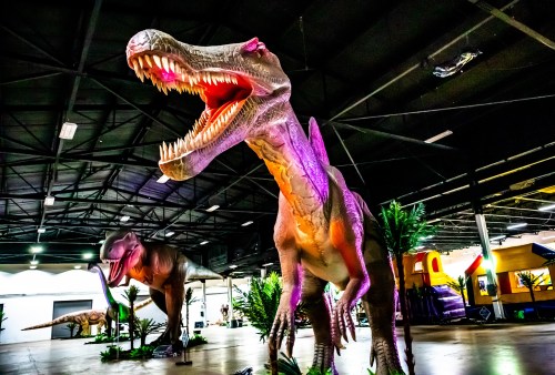 Two large dinosaur sculptures in a dimly lit indoor space, surrounded by artificial plants.