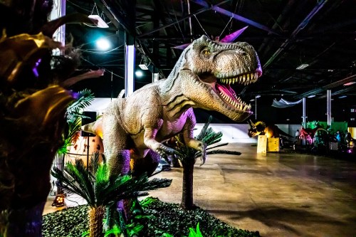 A lifelike dinosaur model stands among palm trees in a dimly lit indoor space, showcasing a prehistoric scene.