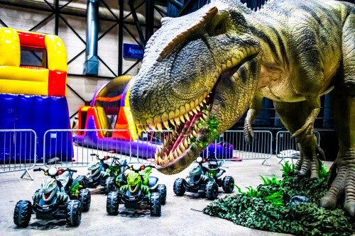 A large dinosaur statue looms over toy quad bikes in a colorful indoor play area with inflatable structures.