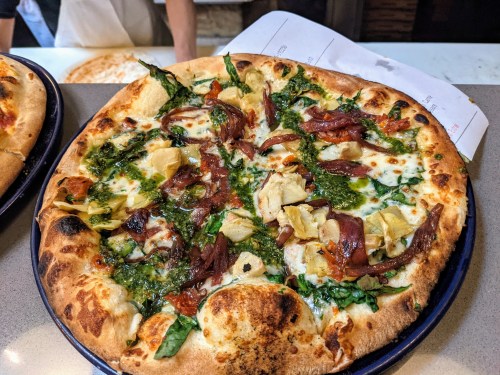 A freshly baked pizza topped with pesto, artichokes, greens, and slices of cured meat on a rustic crust.