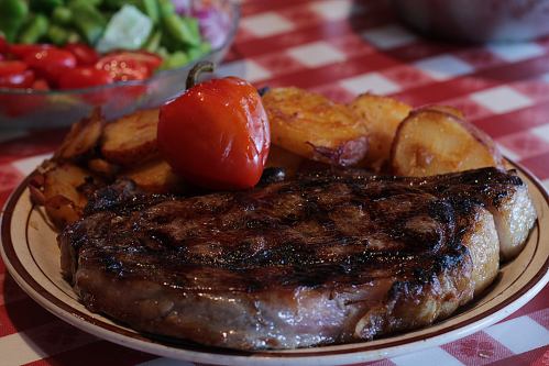 A juicy steak with grilled potatoes and a red pepper, served on a plate with a checkered tablecloth in the background.