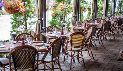 A cozy restaurant with empty tables set for dining, surrounded by greenery and colorful hanging flowers.