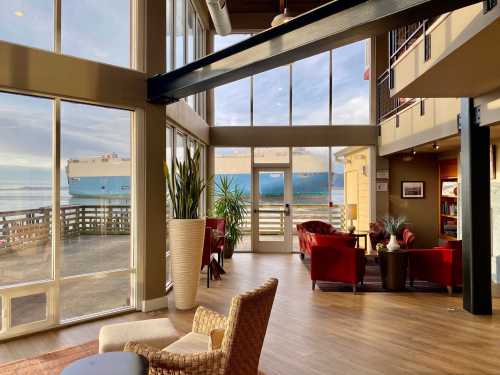 A modern interior with large windows showcasing a view of a ship outside, surrounded by stylish furniture and plants.